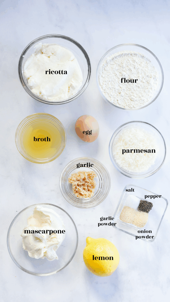 Top-down view of labeled ingredients for gnocchi di ricotta in bowls on a white surface: ricotta, flour, egg, broth, parmesan, garlic, mascarpone, a lemon, plus small bowls of salt, pepper, garlic powder, and onion powder.