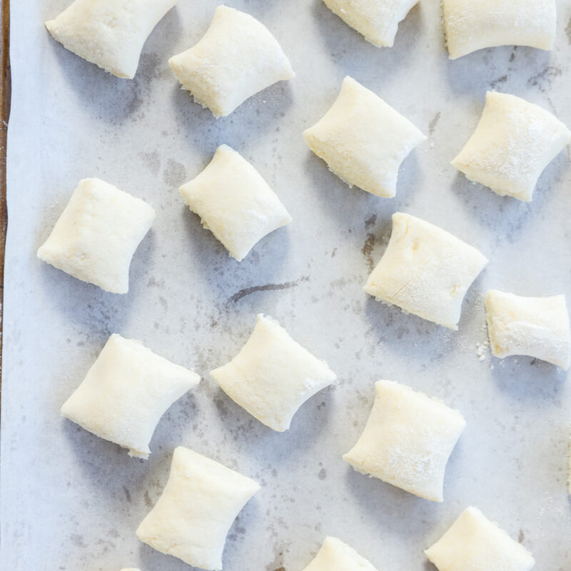 Rows of uncooked gnocchi di ricotta are arranged on a parchment-lined baking sheet, ready for cooking. The delicate gnocchi are lightly dusted with flour and evenly spaced on the tray.