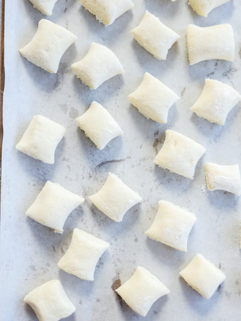 Rows of uncooked gnocchi di ricotta are arranged on a parchment-lined baking sheet, ready for cooking. The delicate gnocchi are lightly dusted with flour and evenly spaced on the tray.