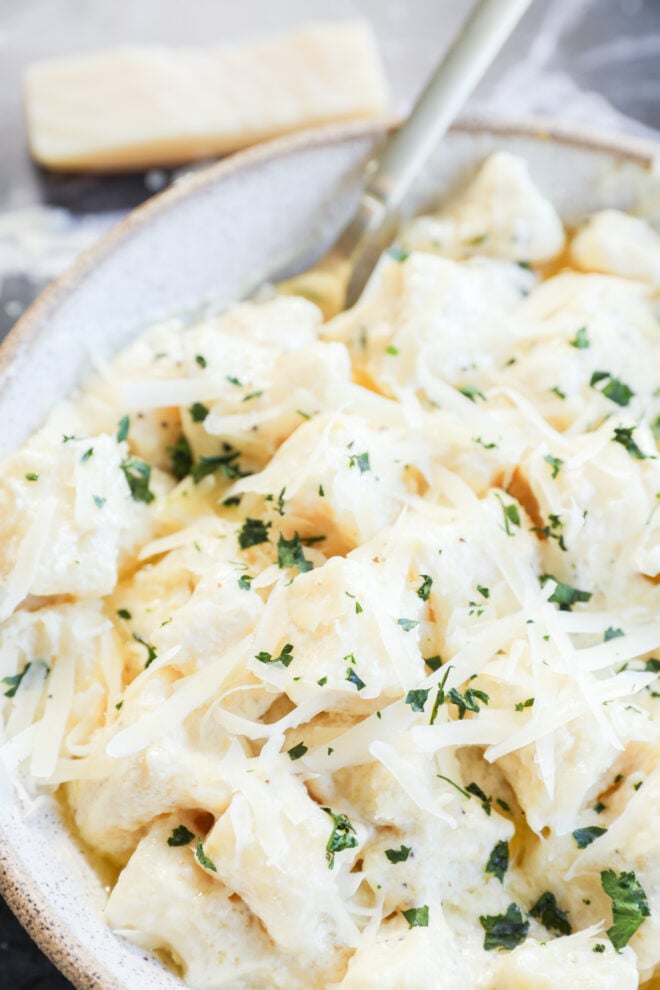 A close-up of creamy gnocchi di ricotta topped with grated cheese and chopped parsley in a bowl, with a fork and a block of cheese in the background.