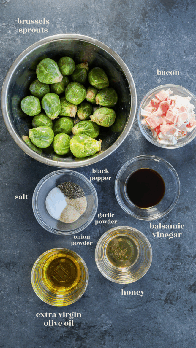 A bowl of raw Brussels sprouts, a bowl of chopped bacon, and small bowls containing salt, black pepper, onion powder, garlic powder, balsamic vinegar, honey, and extra virgin olive oil on a dark surface.