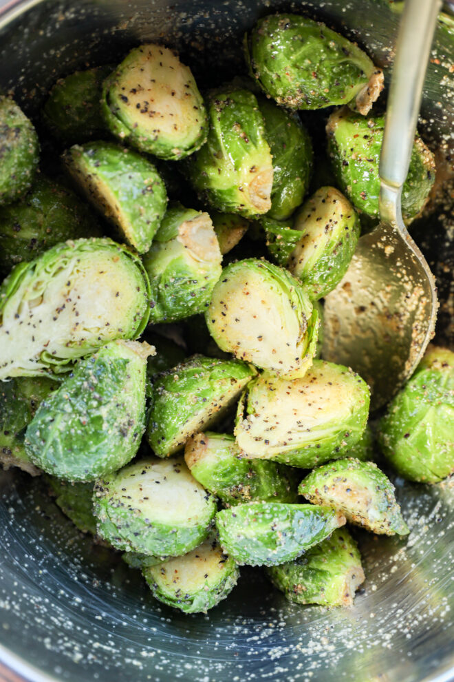 Halved Brussels sprouts coated in seasoning and grated cheese, resting in a metal bowl with a serving spoon.