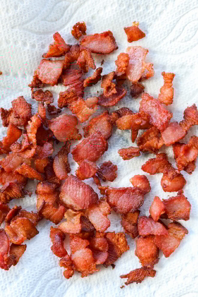 Crispy, cooked bacon pieces scattered on a white paper towel, showing a range of golden brown and red hues, with visible texture and edges.