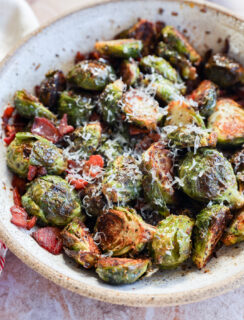 A ceramic bowl filled with roasted Brussels sprouts, topped with grated cheese and pieces of crispy bacon. The sprouts are golden brown and slightly charred, sitting on a light-colored surface.
