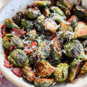 A ceramic bowl filled with roasted Brussels sprouts, topped with grated cheese and pieces of crispy bacon. The sprouts are golden brown and slightly charred, sitting on a light-colored surface.