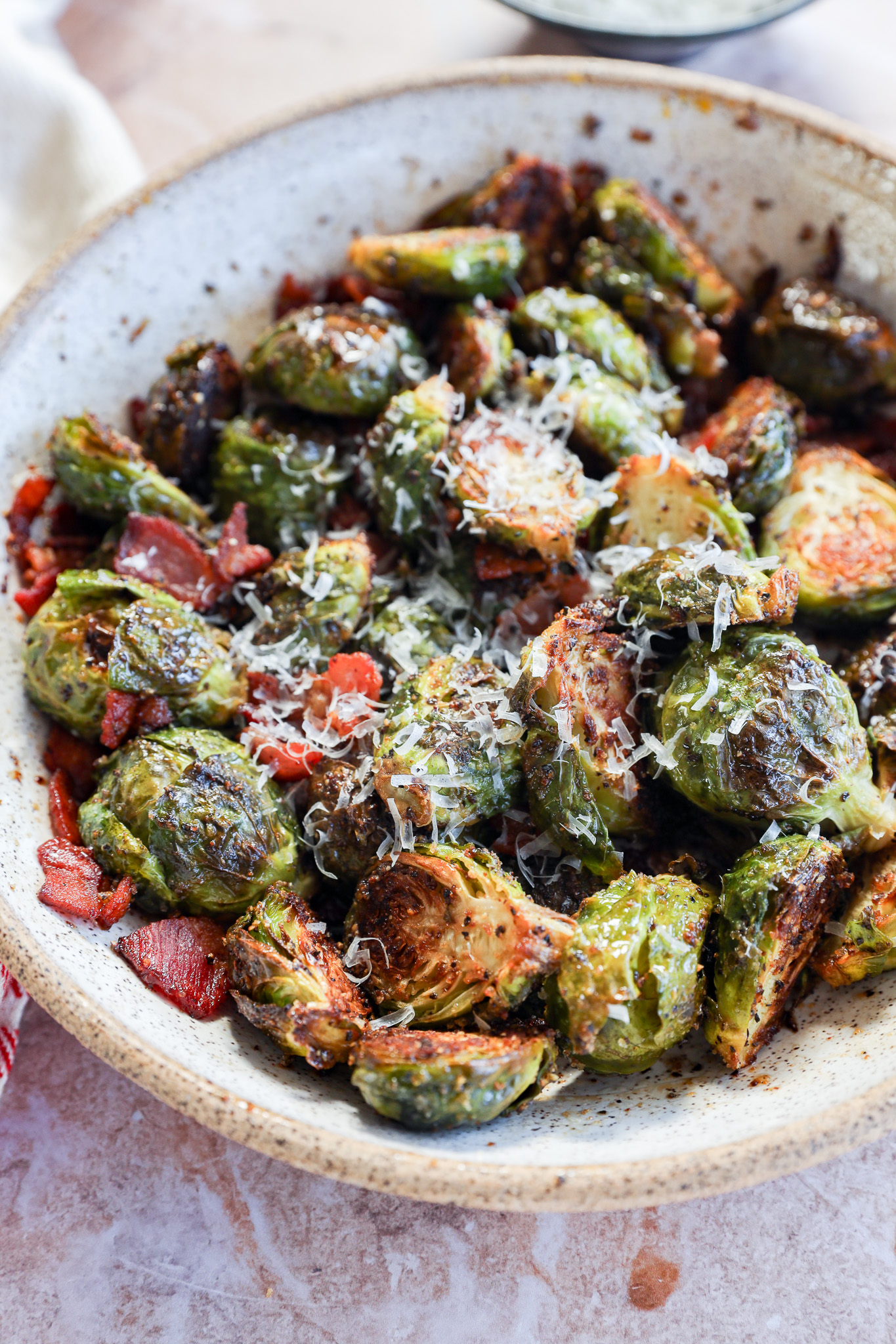 A ceramic bowl filled with roasted Brussels sprouts, topped with grated cheese and pieces of crispy bacon. The sprouts are golden brown and slightly charred, sitting on a light-colored surface.