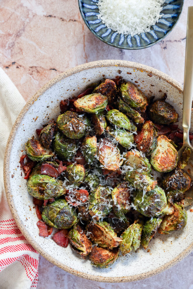 A bowl of roasted Brussels sprouts topped with grated cheese, served with pieces of crispy bacon. A fork rests in the bowl, and a small dish of extra grated cheese is nearby on a marble surface.