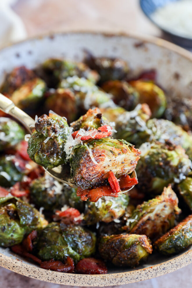 A close-up of a fork holding roasted Brussels sprouts with crispy edges, bacon bits, and grated cheese, above a bowl filled with more of the same dish.