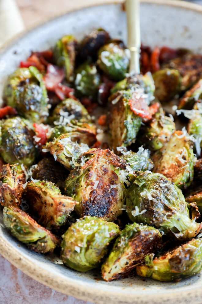 A bowl of roasted Brussels sprouts garnished with grated cheese and small bacon pieces, with a fork resting inside the dish. The sprouts are browned and crispy.