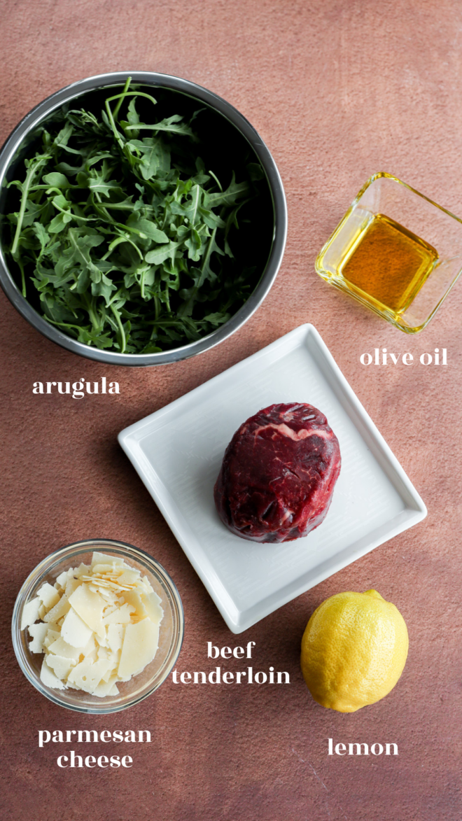 Five ingredients for Carpaccio di Manzo are arranged on a brown surface: arugula in a bowl, olive oil in a small glass, beef tenderloin on a white plate, parmesan cheese shavings in a bowl, and a whole lemon. Each is labeled.