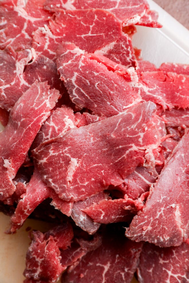 Thin slices of raw marbled beef, known as Carpaccio di Manzo, are stacked on a white surface, showcasing a bright red color with visible streaks of white fat.