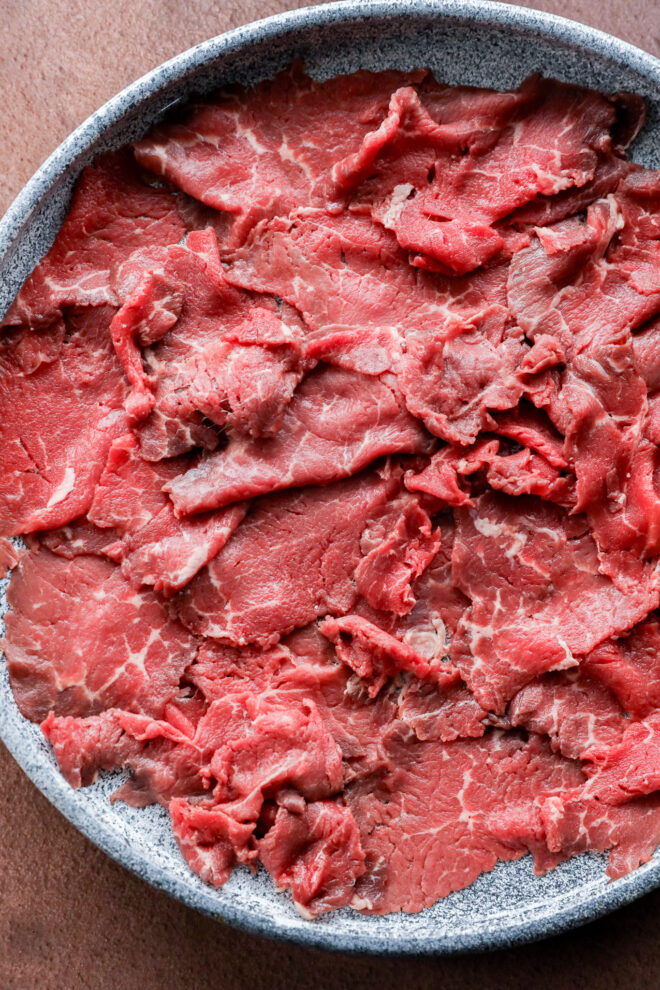A close-up of thinly sliced raw beef, layered on a round gray plate—perfect for cooking or use in dishes like hot pot or stir-fry.