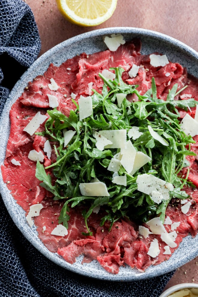A plate of thinly sliced meat topped with fresh arugula and shaved Parmesan cheese, served on a gray dish with a lemon wedge on the side.