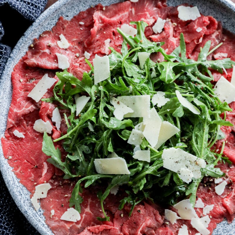 A plate of thinly sliced meat topped with fresh arugula and shaved Parmesan cheese, served on a gray dish with a lemon wedge on the side.