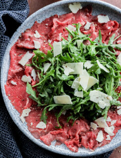 A plate of Carpaccio di Manzo features thinly sliced raw beef topped with a mound of arugula and shaved parmesan cheese, garnished with cracked black pepper. A navy cloth and lemon wedge are nearby.