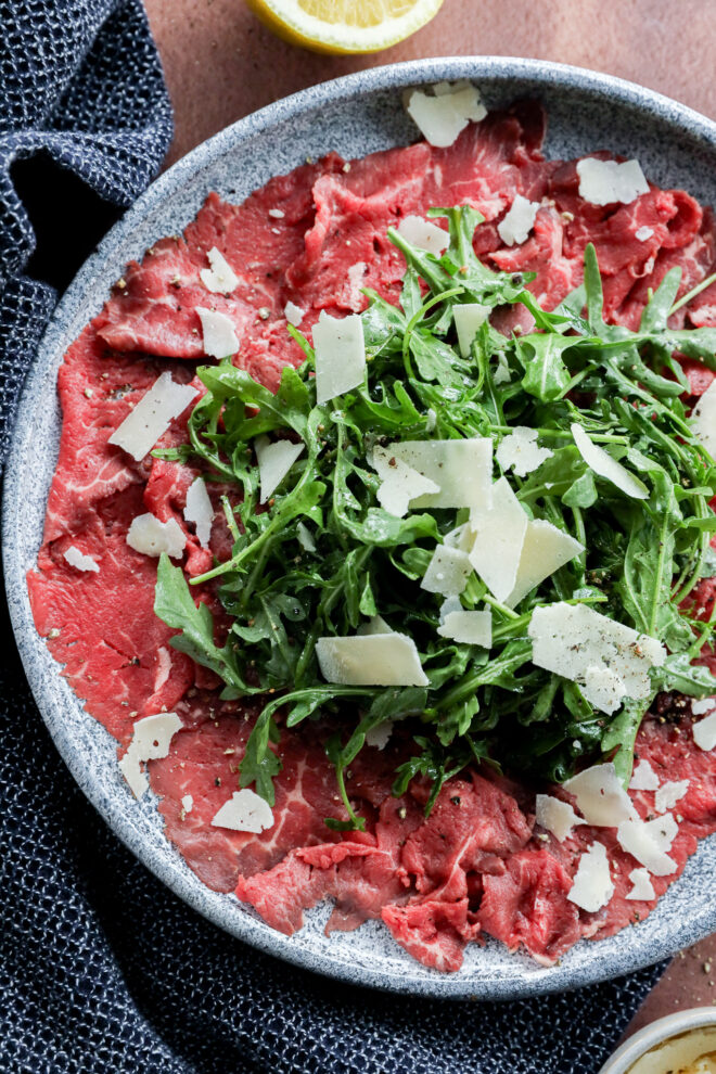 A plate of Carpaccio di Manzo features thinly sliced raw beef topped with a mound of arugula and shaved parmesan cheese, garnished with cracked black pepper. A navy cloth and lemon wedge are nearby.
