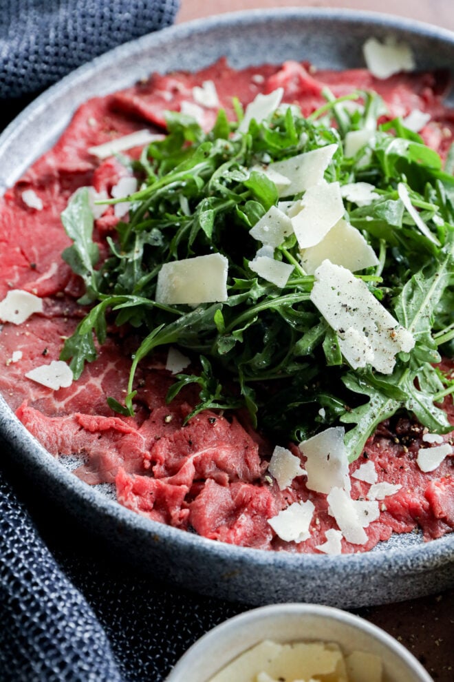 A plate of Carpaccio di Manzo features thinly sliced raw beef topped with arugula, shaved Parmesan cheese, and ground black pepper.