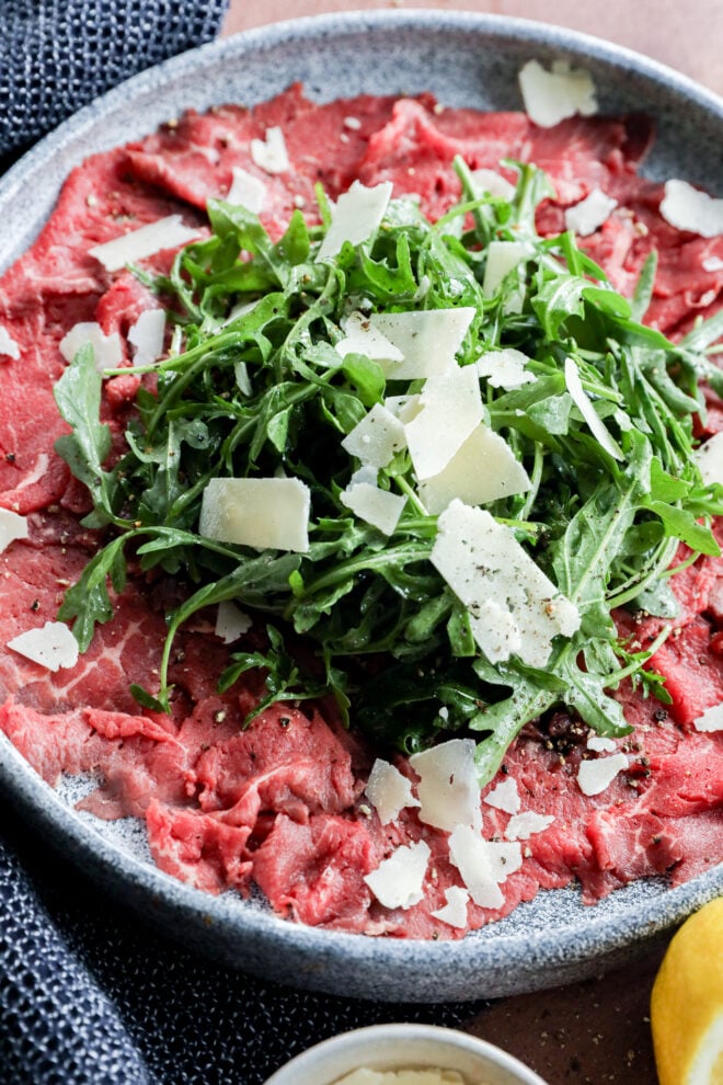 A plate of beef topped with a mound of fresh arugula and shaved Parmesan cheese, garnished with cracked black pepper.