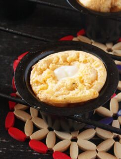 A small, golden-brown cornbread with a pat of melting butter in the center sits in a black cast iron skillet, placed on a decorative trivet with red, beige, and blue accents.