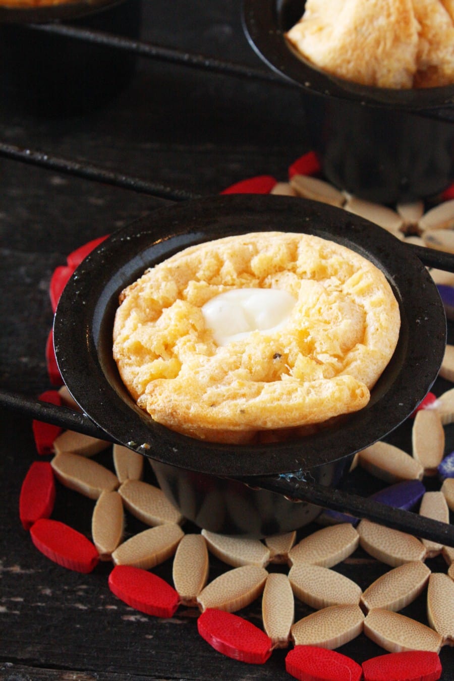 A small, golden-brown cornbread with a pat of melting butter in the center sits in a black cast iron skillet, placed on a decorative trivet with red, beige, and blue accents.