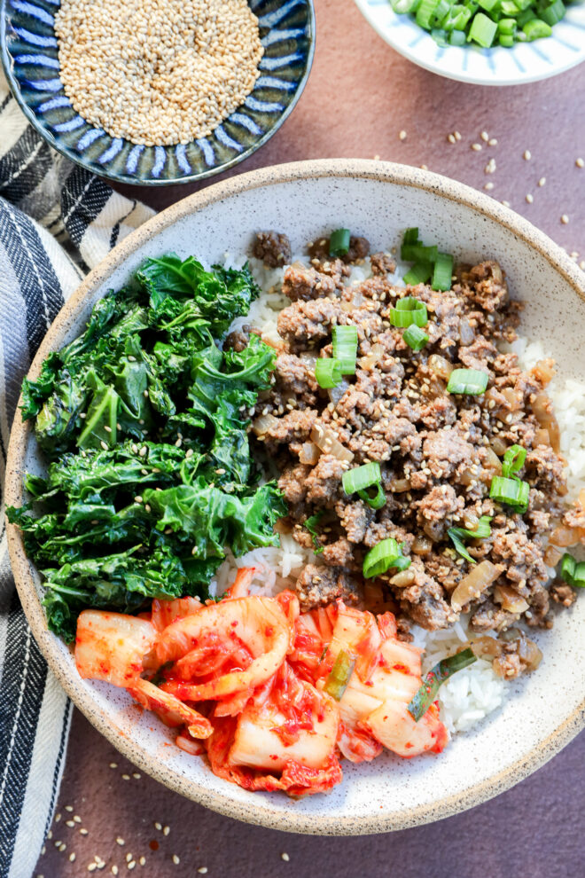 A bowl of white rice topped with cooked ground beef and chopped green onions, sautéed kale, and a serving of kimchi. Bowls of sesame seeds and chopped green onions sit nearby on a brown surface.