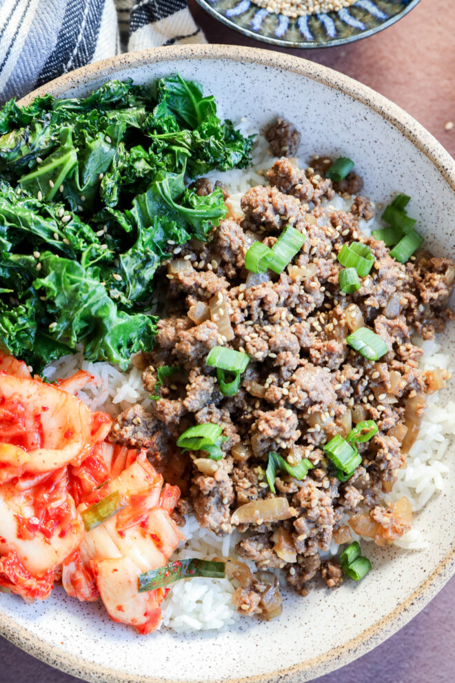 A bowl filled with white rice, topped with ground beef and chopped green onions, sautéed kale, and a serving of kimchi.