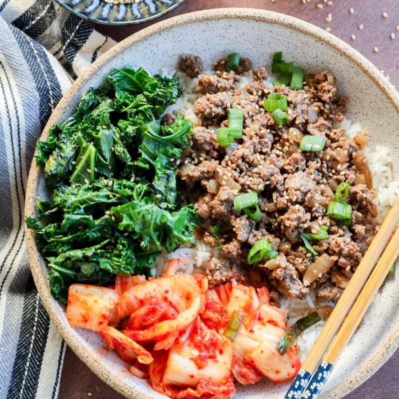 A bowl of white rice topped with cooked ground beef, saut&eacute;ed greens, and kimchi, garnished with chopped green onions. Chopsticks rest on the bowl, with small bowls of sesame seeds and green onions nearby.