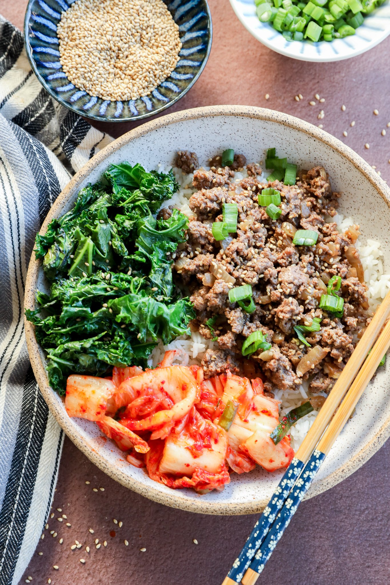 A bowl of white rice topped with cooked ground beef, sautéed greens, and kimchi, garnished with chopped green onions. Chopsticks rest on the bowl, with small bowls of sesame seeds and green onions nearby.