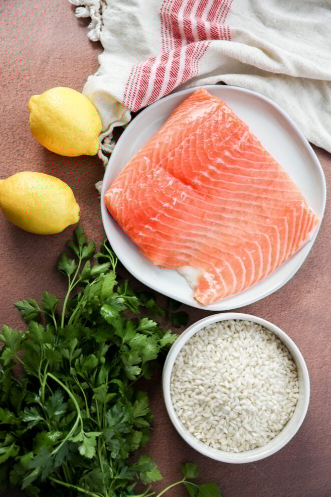 A raw salmon fillet on a white plate, two lemons, fresh parsley, and a bowl of uncooked rice are arranged on a brown surface.
