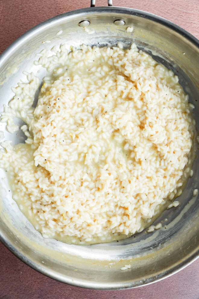 A close-up of creamy risotto in a stainless steel pan, showcasing a smooth, slightly glossy texture and light color, sitting on a brown surface.