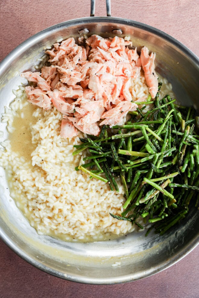 A stainless steel pan containing cooked rice, flaked salmon, and saut&eacute;ed asparagus pieces&mdash;reminiscent of a deconstructed Salmon Lemon Risotto with Asparagus&mdash;each ingredient separated into sections on a pink surface.