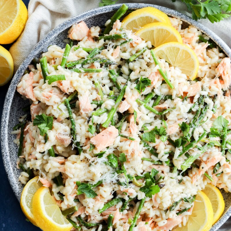 A gray bowl filled with Salmon Lemon Risotto with Asparagus, creamy risotto mixed with salmon, asparagus, and fresh herbs, garnished with grated cheese and lemon wedges. A bunch of cilantro, a white napkin, and lemon halves are nearby on a blue surface.