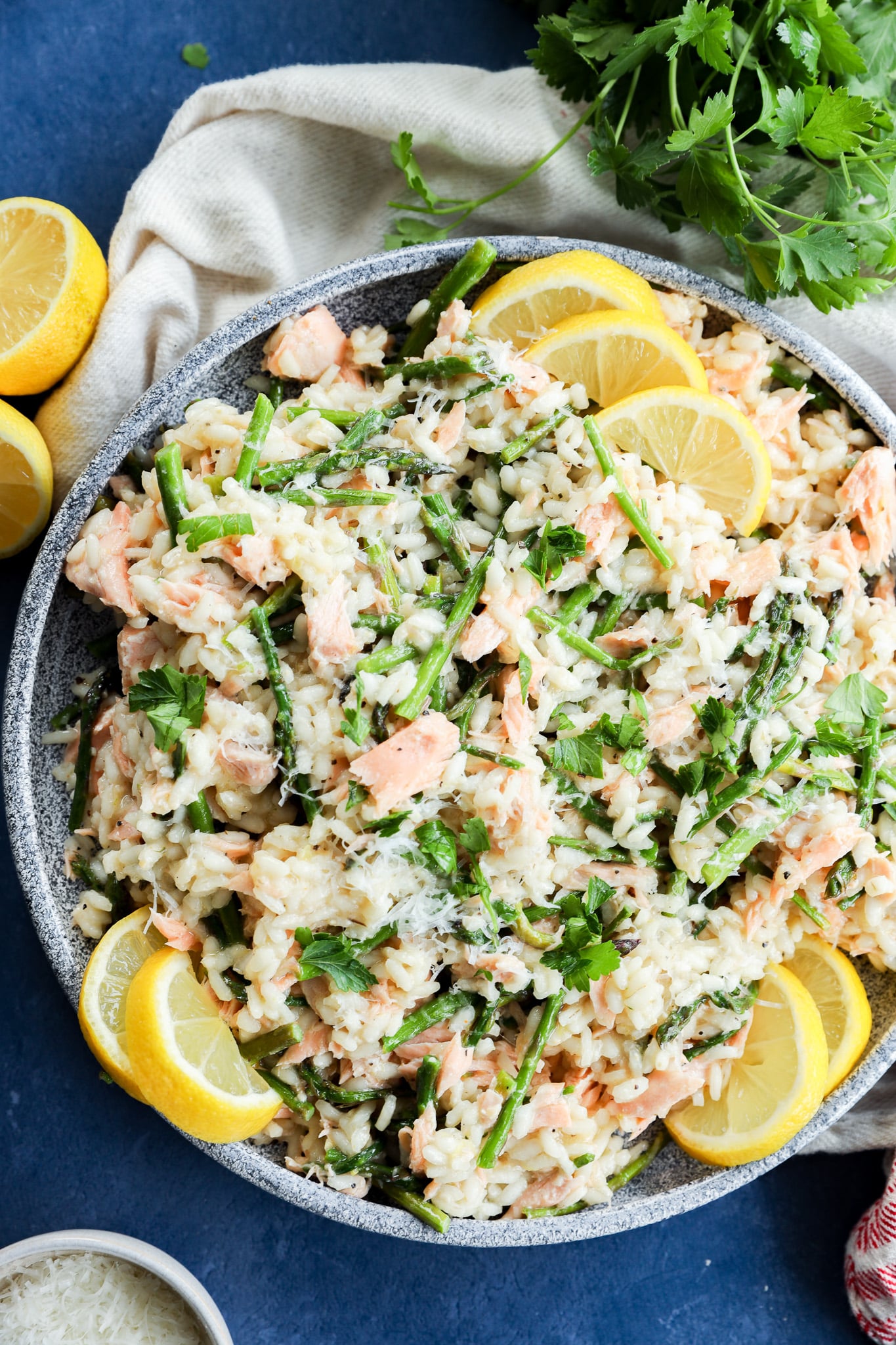 A gray bowl filled with Salmon Lemon Risotto with Asparagus, creamy risotto mixed with salmon, asparagus, and fresh herbs, garnished with grated cheese and lemon wedges. A bunch of cilantro, a white napkin, and lemon halves are nearby on a blue surface.