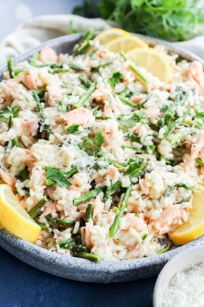 A close-up of a bowl of Salmon Lemon Risotto with Asparagus, featuring creamy risotto, salmon pieces, fresh herbs, grated cheese, and lemon wedges. The dish is colorful and looks fresh and appetizing.