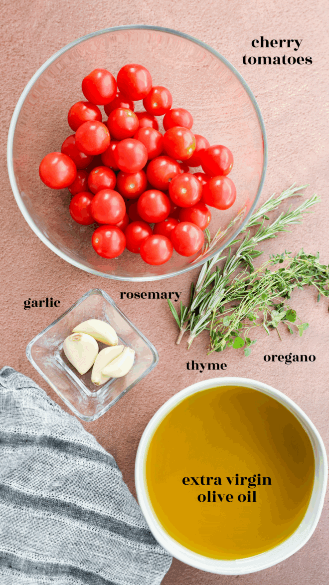 A bowl of cherry tomatoes, perfect for making cherry tomato confit, sits with extra virgin olive oil, garlic cloves, and sprigs of rosemary, thyme, and oregano on a pink surface next to a gray striped cloth. Ingredients are clearly labeled.