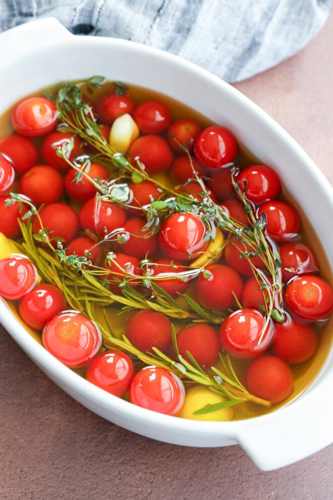 A white dish filled with cherry tomato confit, sprigs of fresh rosemary and thyme, and garlic cloves, all submerged in olive oil, before cooking, with a gray cloth in the background.