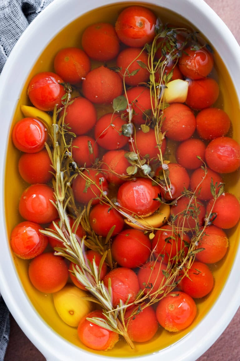Cherry tomato confit is made by submerging cherry tomatoes, garlic cloves, and fresh herbs like rosemary and thyme in olive oil in a white oval dish.