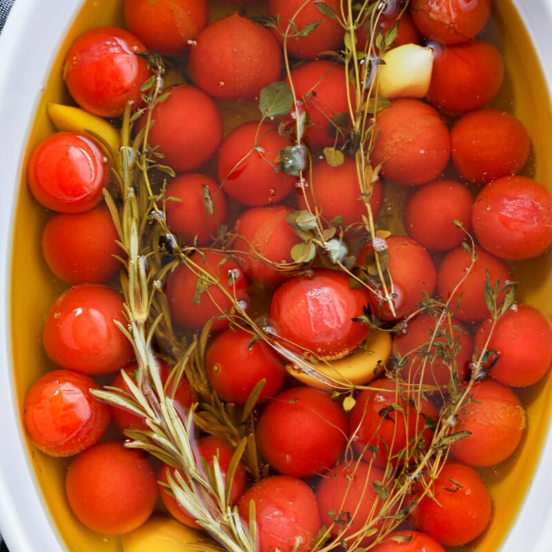 Cherry tomato confit is made by submerging cherry tomatoes, garlic cloves, and fresh herbs like rosemary and thyme in olive oil in a white oval dish.