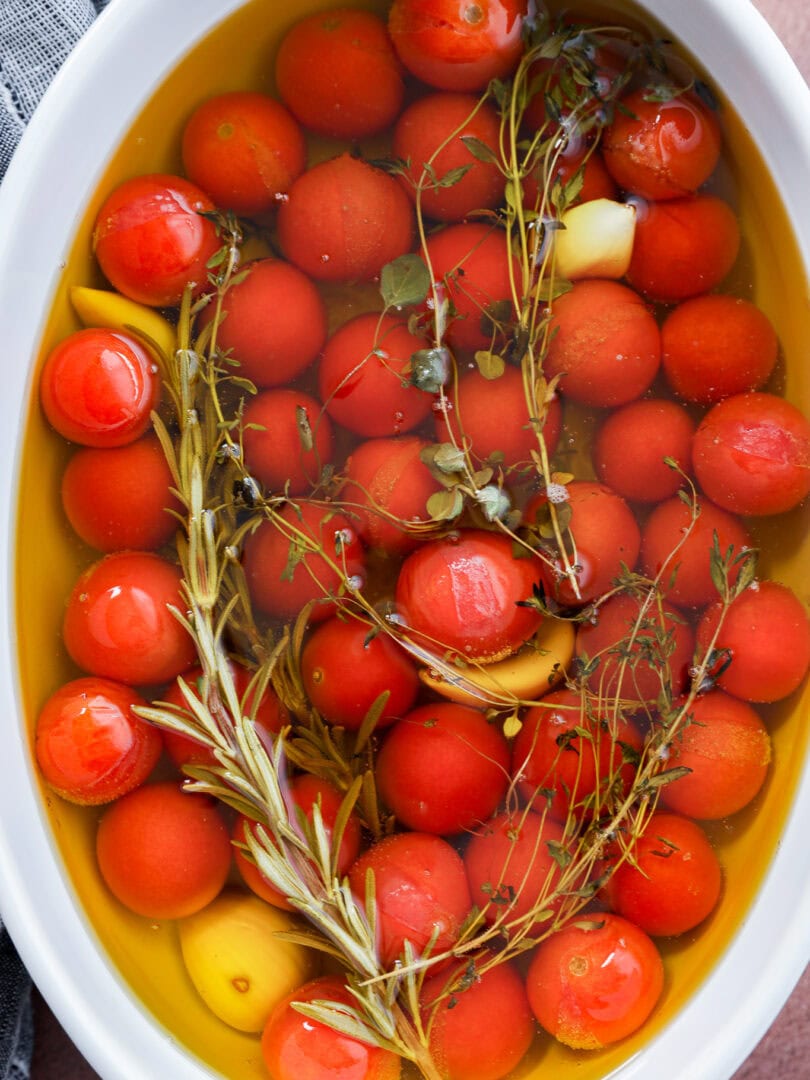 Cherry tomato confit is made by submerging cherry tomatoes, garlic cloves, and fresh herbs like rosemary and thyme in olive oil in a white oval dish.