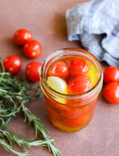A jar of cherry tomato confit—cherry tomatoes, olive oil, and a clove of garlic—rests on a brown surface beside fresh rosemary, loose cherry tomatoes, and a blue checkered cloth.