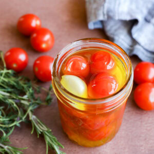 A jar of cherry tomato confit—cherry tomatoes, olive oil, and a clove of garlic—rests on a brown surface beside fresh rosemary, loose cherry tomatoes, and a blue checkered cloth.