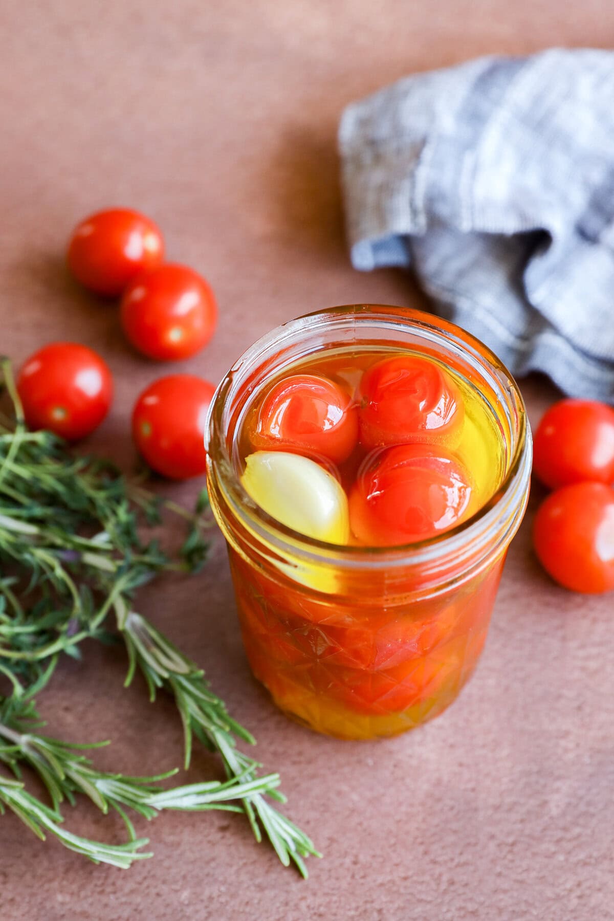 A jar of cherry tomato confit—cherry tomatoes, olive oil, and a clove of garlic—rests on a brown surface beside fresh rosemary, loose cherry tomatoes, and a blue checkered cloth.