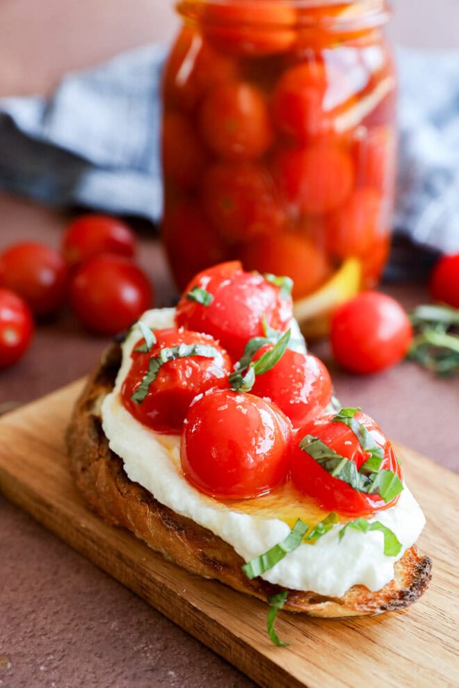 A slice of toasted bread topped with creamy cheese, cherry tomatoes, and fresh basil sits on a wooden board. In the background, a jar filled with more cherry tomatoes is visible.