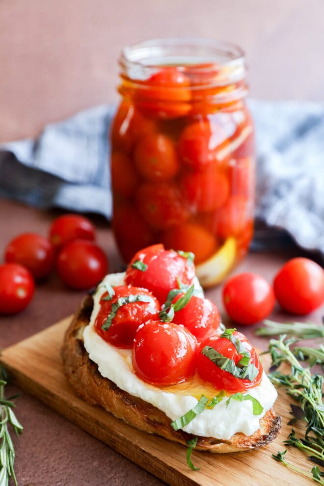 A slice of toasted bread topped with creamy cheese, cherry tomato confit, and fresh basil sits on a wooden board. In the background, a jar filled with cherry tomatoes and a few loose tomatoes are visible.