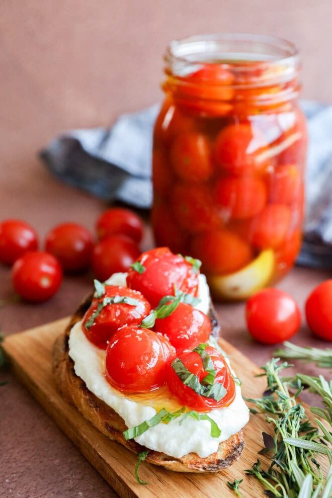 A slice of toasted bread topped with fresh mozzarella, cherry tomatoes, and basil sits on a wooden board. In the background, a jar of cherry tomato confit and scattered fresh tomatoes and herbs add vibrant color to the scene.
