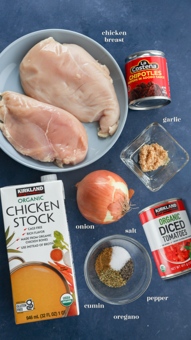 Ingredients for chicken tinga tacos are displayed: raw chicken breasts, canned chipotle peppers, minced garlic, a yellow onion, canned diced tomatoes, organic chicken stock, and small bowls of salt, pepper, cumin, and oregano.