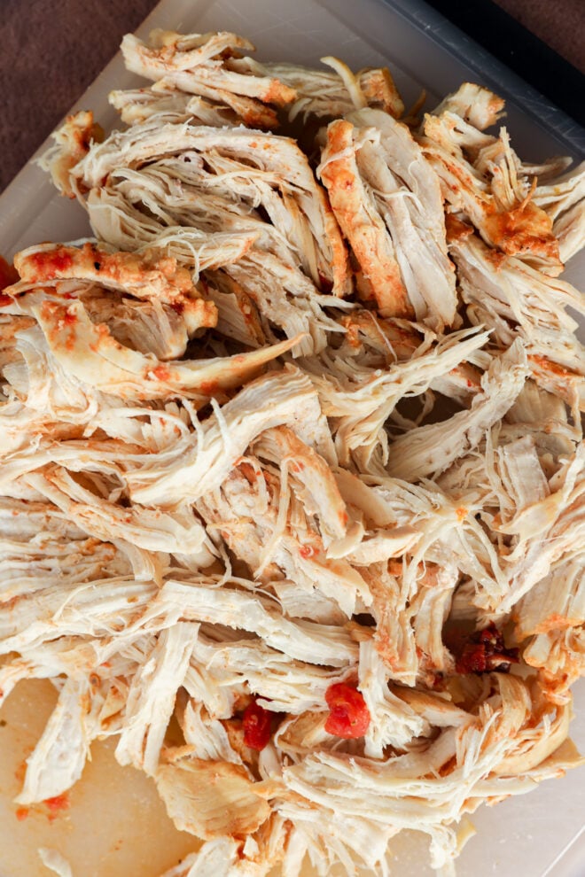 Shredded cooked chicken with a light seasoning and small bits of tomato spread out on a cutting board.