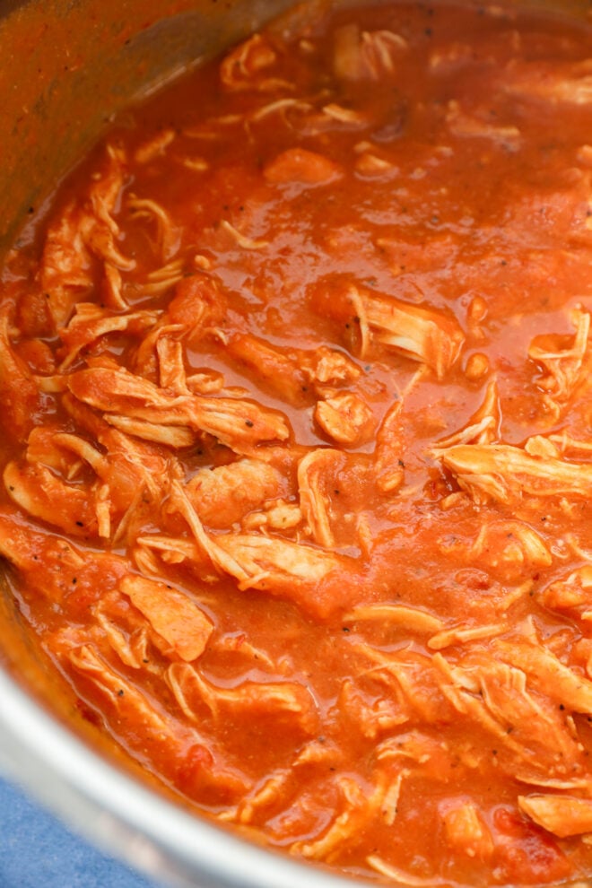 Shredded chicken tinga tacos filling covered in a rich, red tomato-based sauce inside a metal pot.