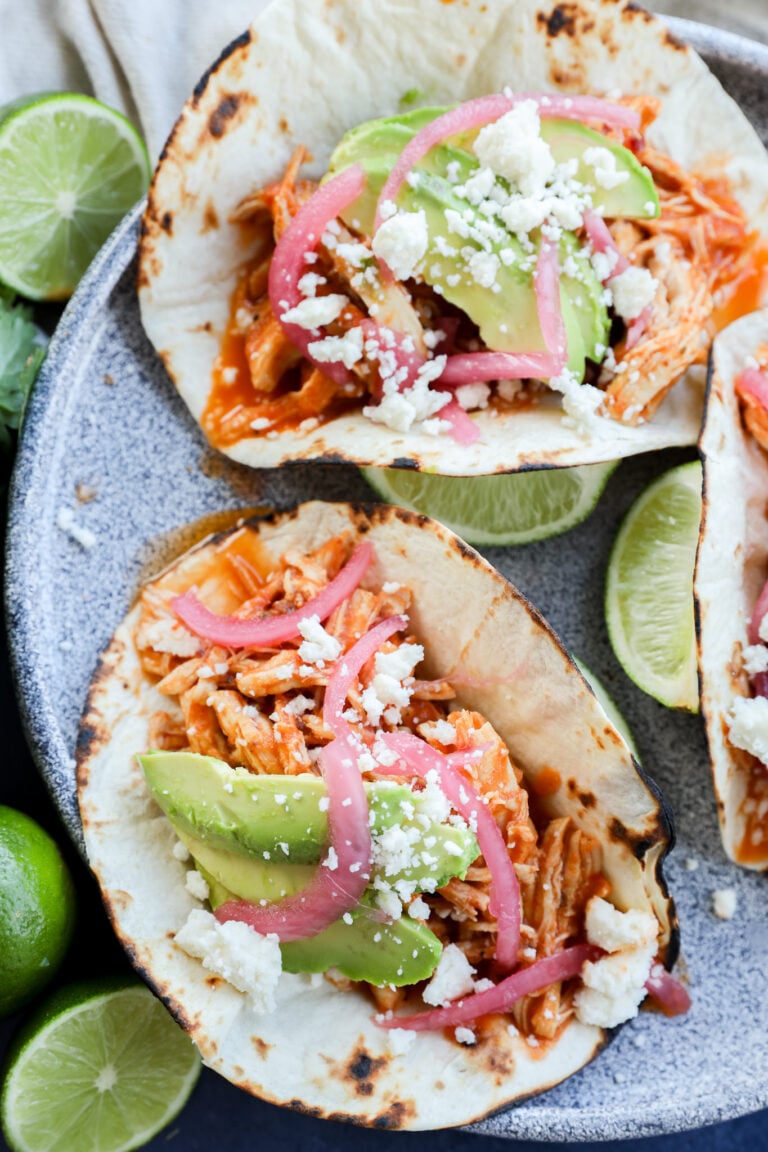 Three chicken tacos on grilled tortillas, topped with sliced avocado, pickled red onions, crumbled cheese, and served with lime wedges on a gray plate.