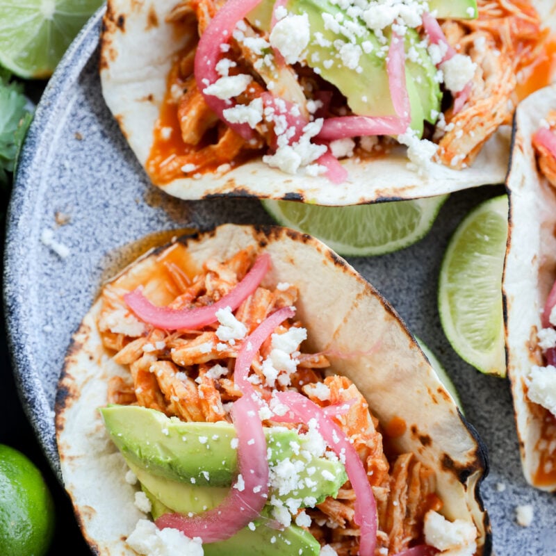 Three chicken tacos on grilled tortillas, topped with sliced avocado, pickled red onions, crumbled cheese, and served with lime wedges on a gray plate.